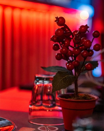 Bacche rosse in un piccolo vaso su un tavolo, con bicchieri e luci rosse sullo sfondo.