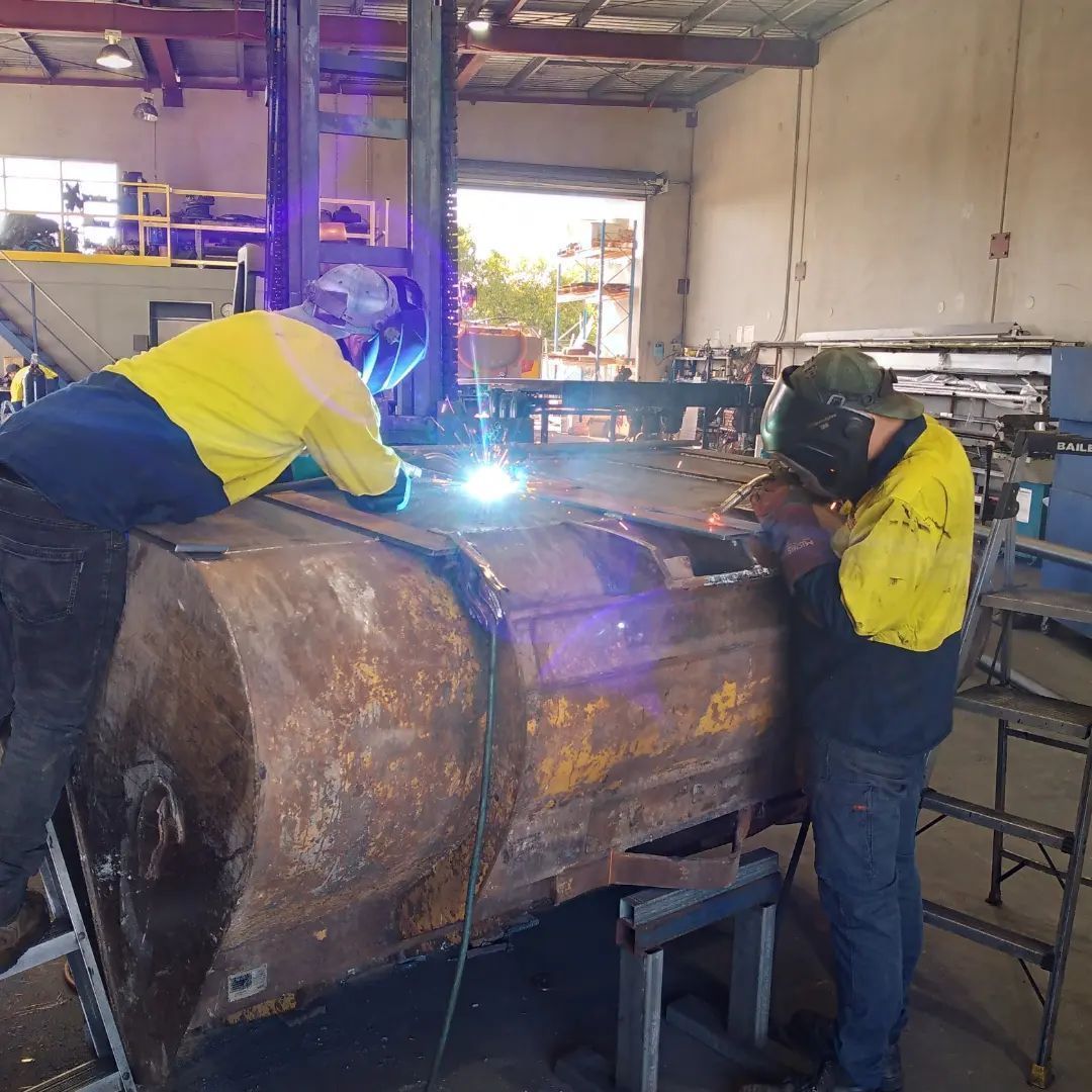 Two welders in yellow shirts working on a large metal structure in a workshop.