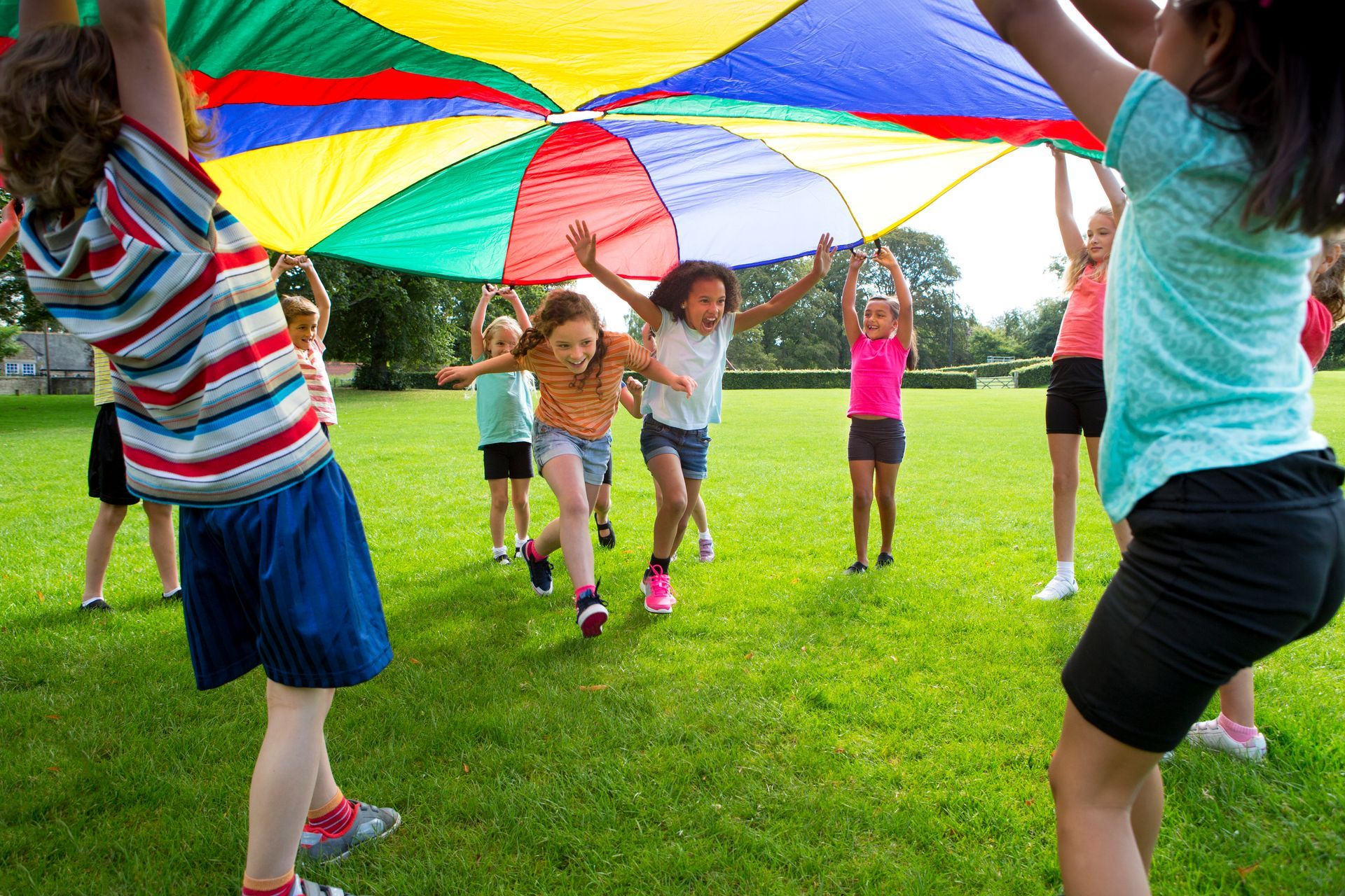 Children in a park holding a colorful parachute with some children running under it.