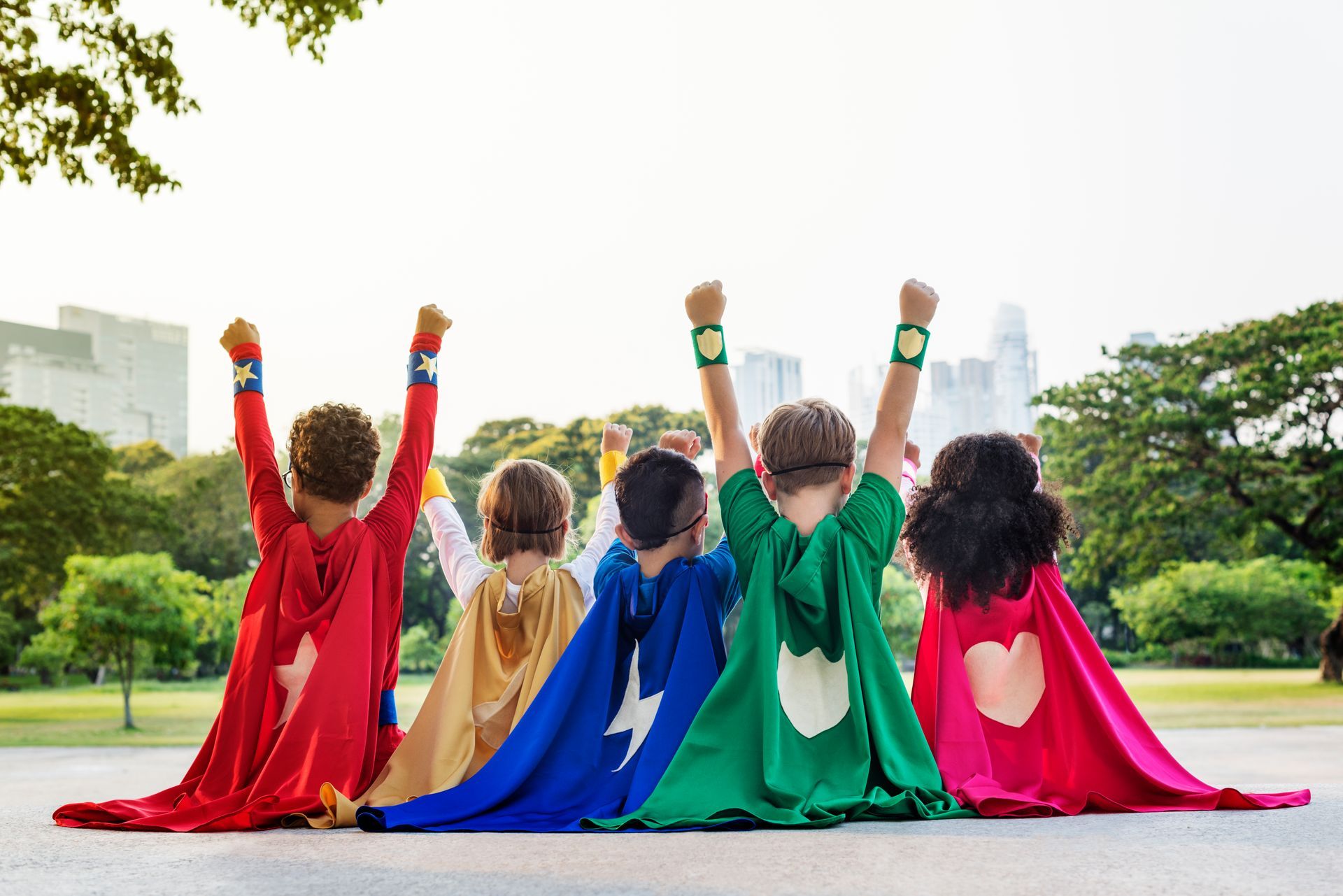 That backs of children sitting down outside with colorful capes on. They have their hands in the air, with trees and sky in the background.