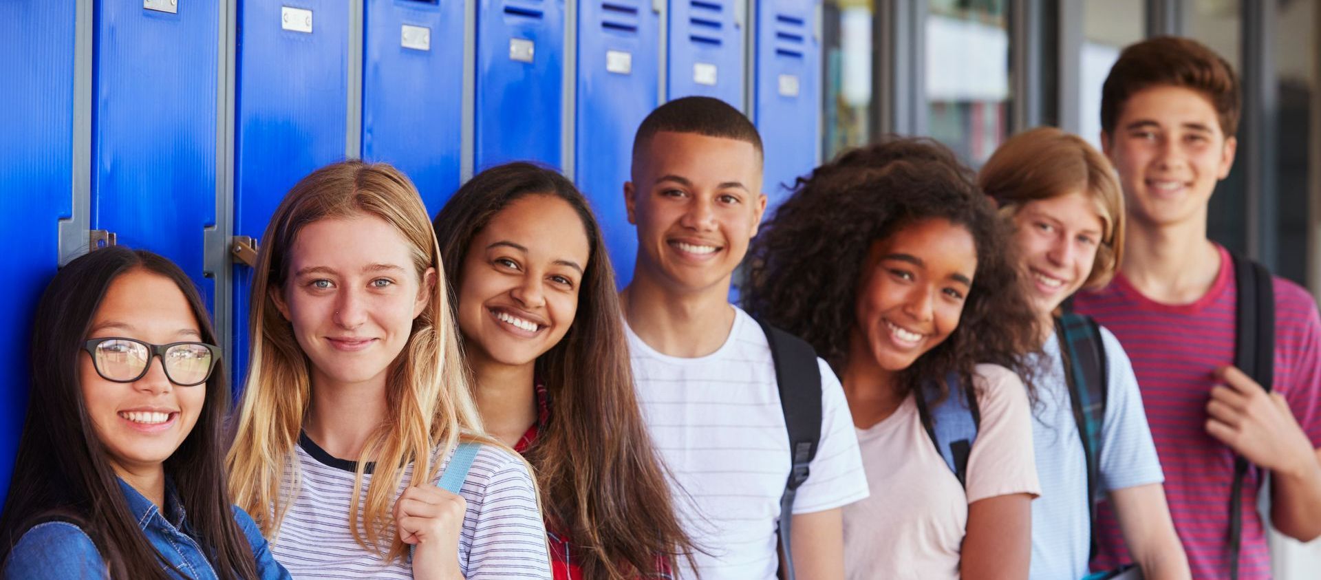 A group of teenagers in front of blue lockers. They are wearing backpacks and smiling.