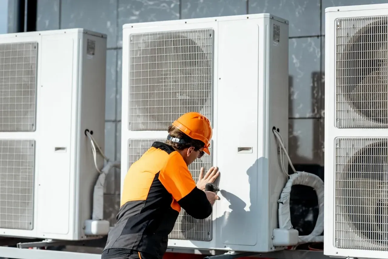 HVAC technician in orange workwear inspecting an outdoor AC unit. Outside, near other units. — Paul Wiedman Electrical in Harristown, QLD