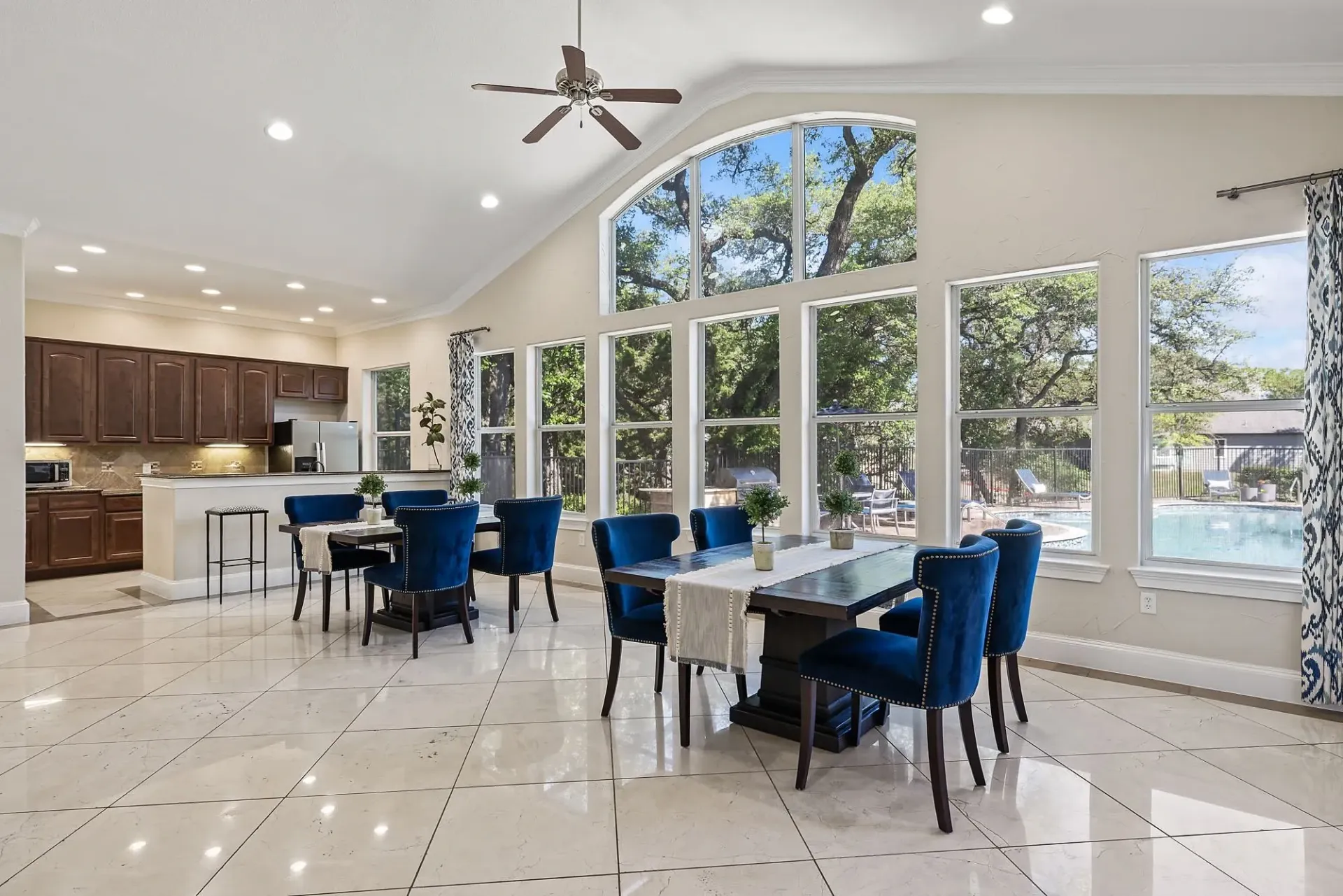 Elegant dining area with plush blue chairs overlooking a sparkling swimming pool and lush green trees.