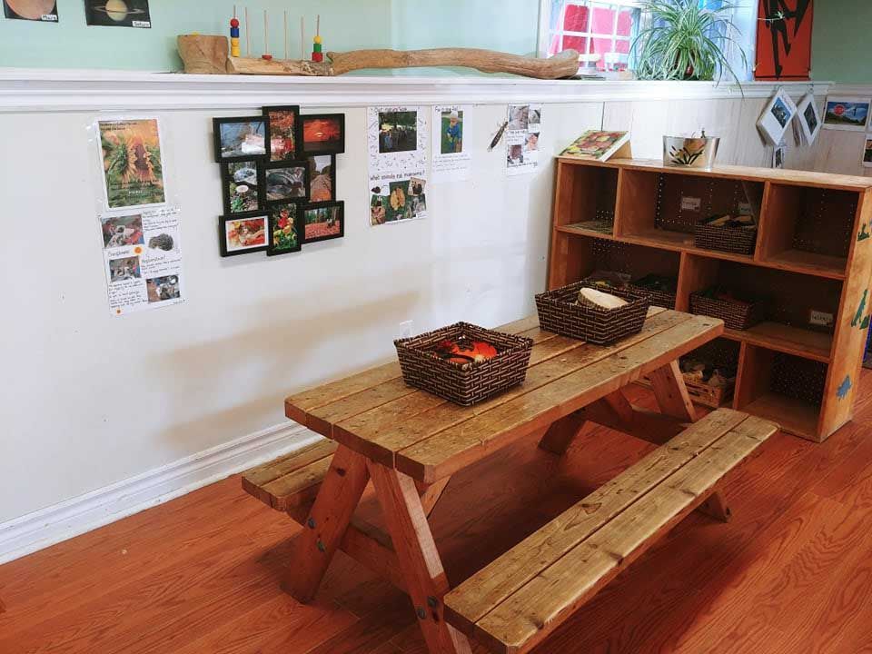 A wooden picnic table with two baskets sits in a brightly lit room, next to a wooden shelf and a white wall with framed photos and educational materials.