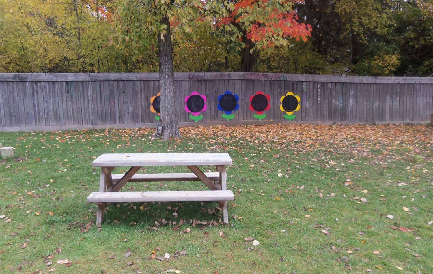 A wooden picnic table sits in a grassy yard with a wooden fence decorated with colorful flower cutouts. Autumn foliage is visible in the background.