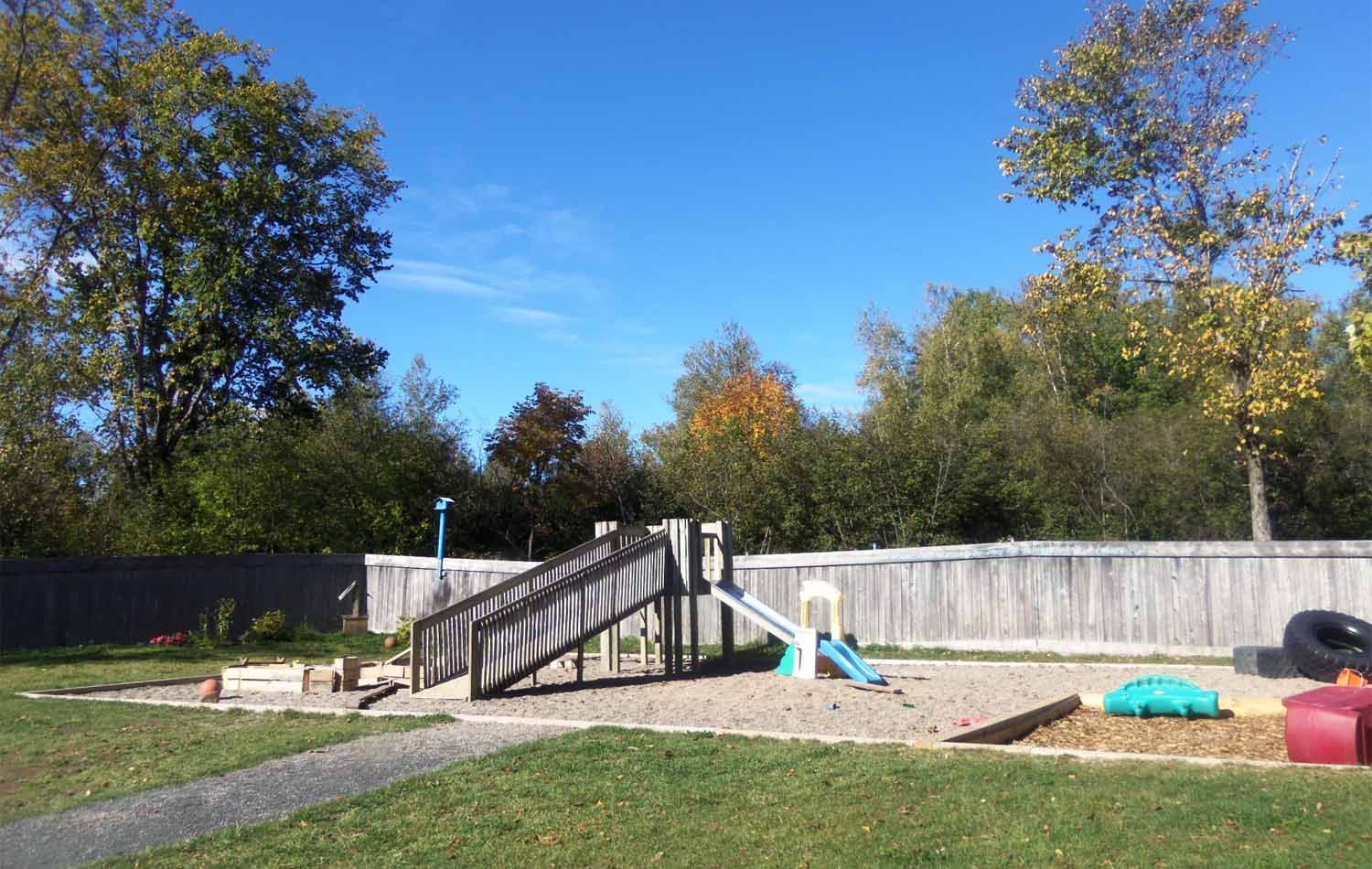 Playground with wooden structure, slide, and sand area, surrounded by a wooden fence, trees, and a blue sky.