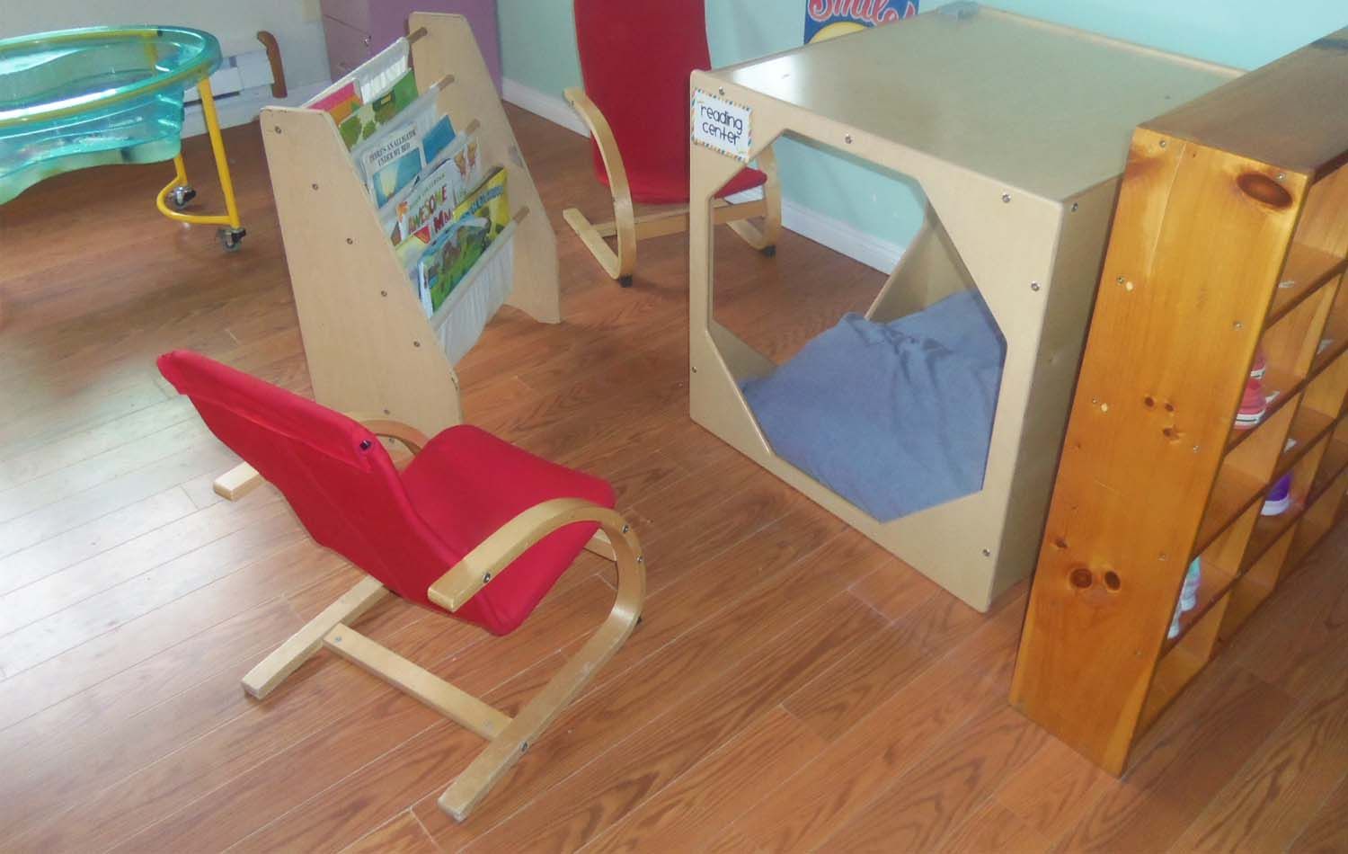 A small playroom with a red chair, bookshelf, reading stand with a book, and a play cubby with a blue mat.
