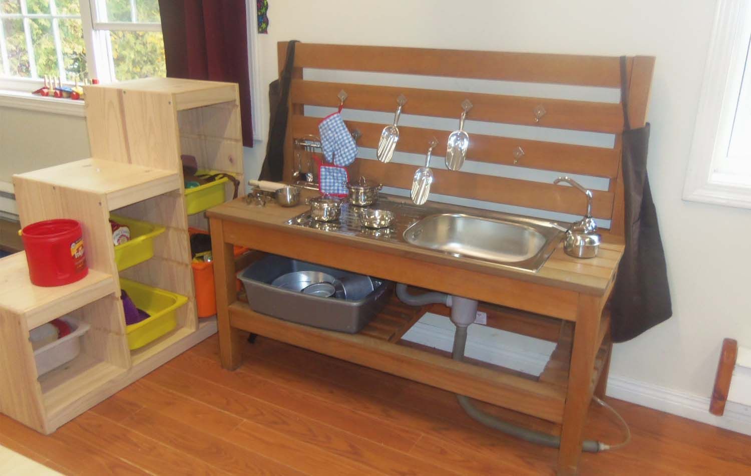 Child's play kitchen with a sink, utensils, and storage on a wooden table, near a window and shelving unit.