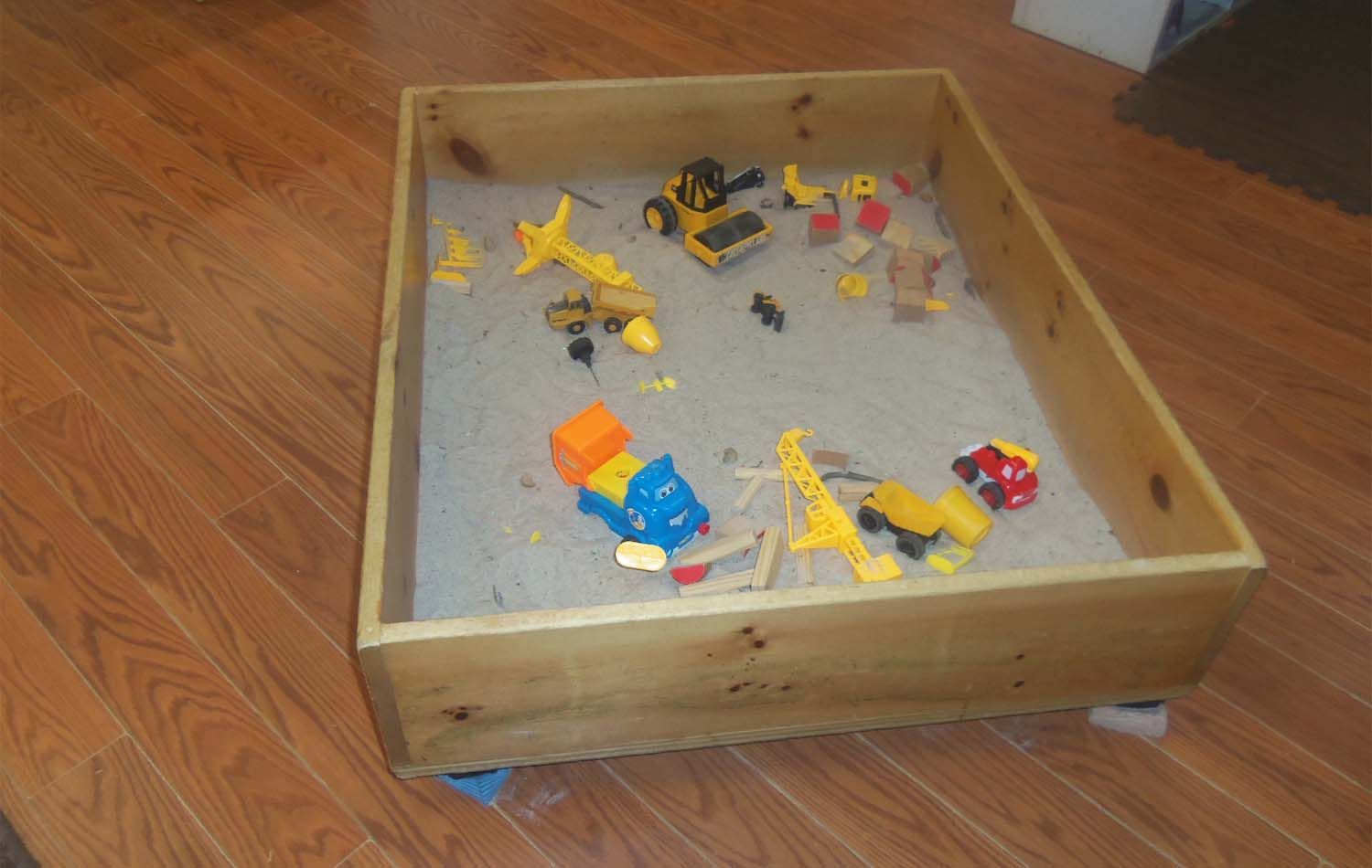 Wooden sandbox filled with sand and construction toys on a wooden floor.