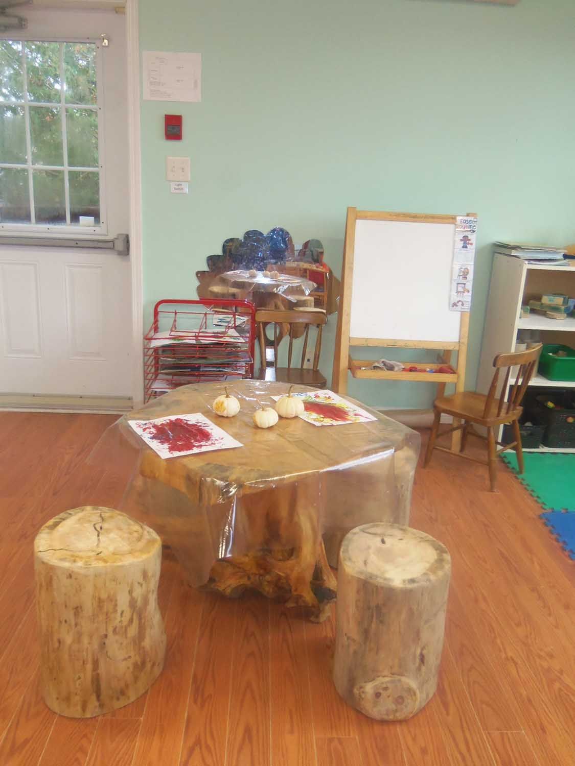 A child's art space: a tree stump table with small log stools, a red cart, and an easel, with pumpkins and art supplies.