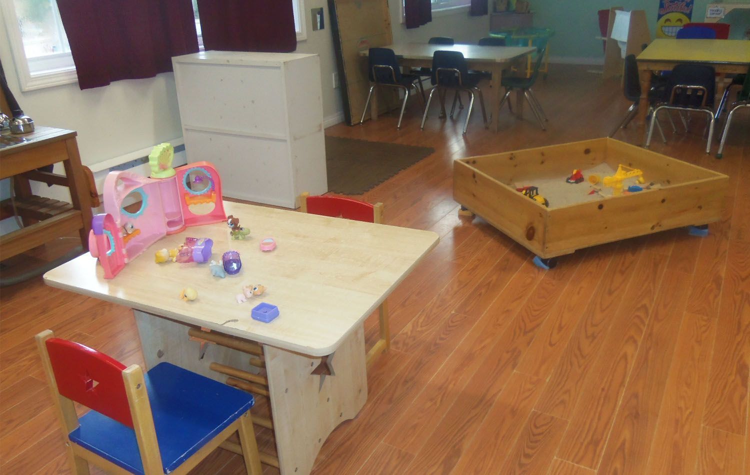 A child's playroom with a wooden table and toys, a sandbox, and small chairs, with a white cabinet in the background.