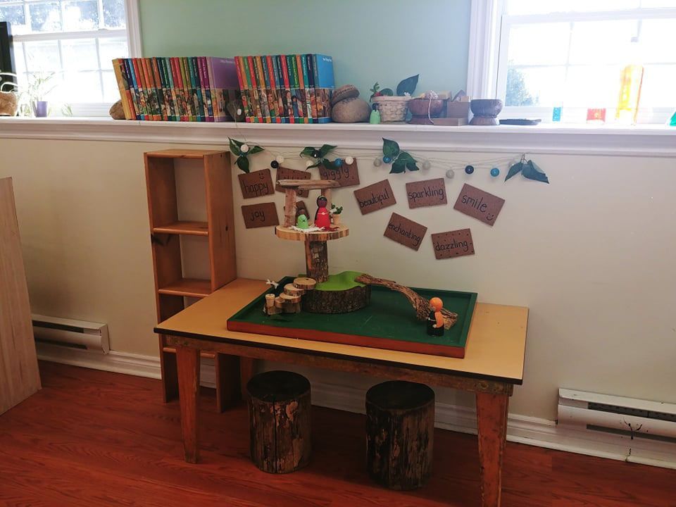 A child-sized table with a forest diorama. Two tree stump seats sit below. A shelf and a window are in the background.