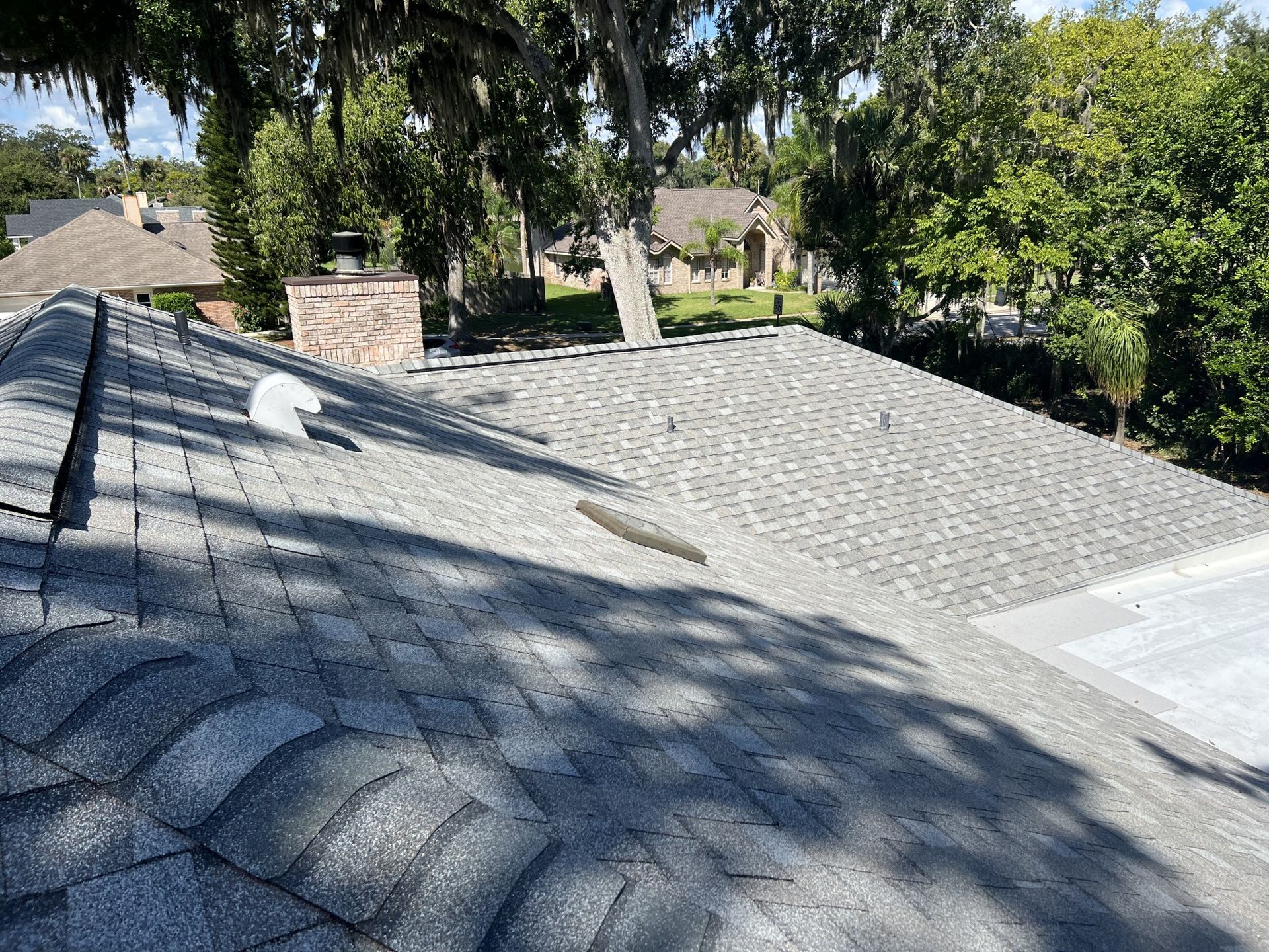 Gray shingled roof with tree shadows and nearby chimneys in a suburban neighborhood
