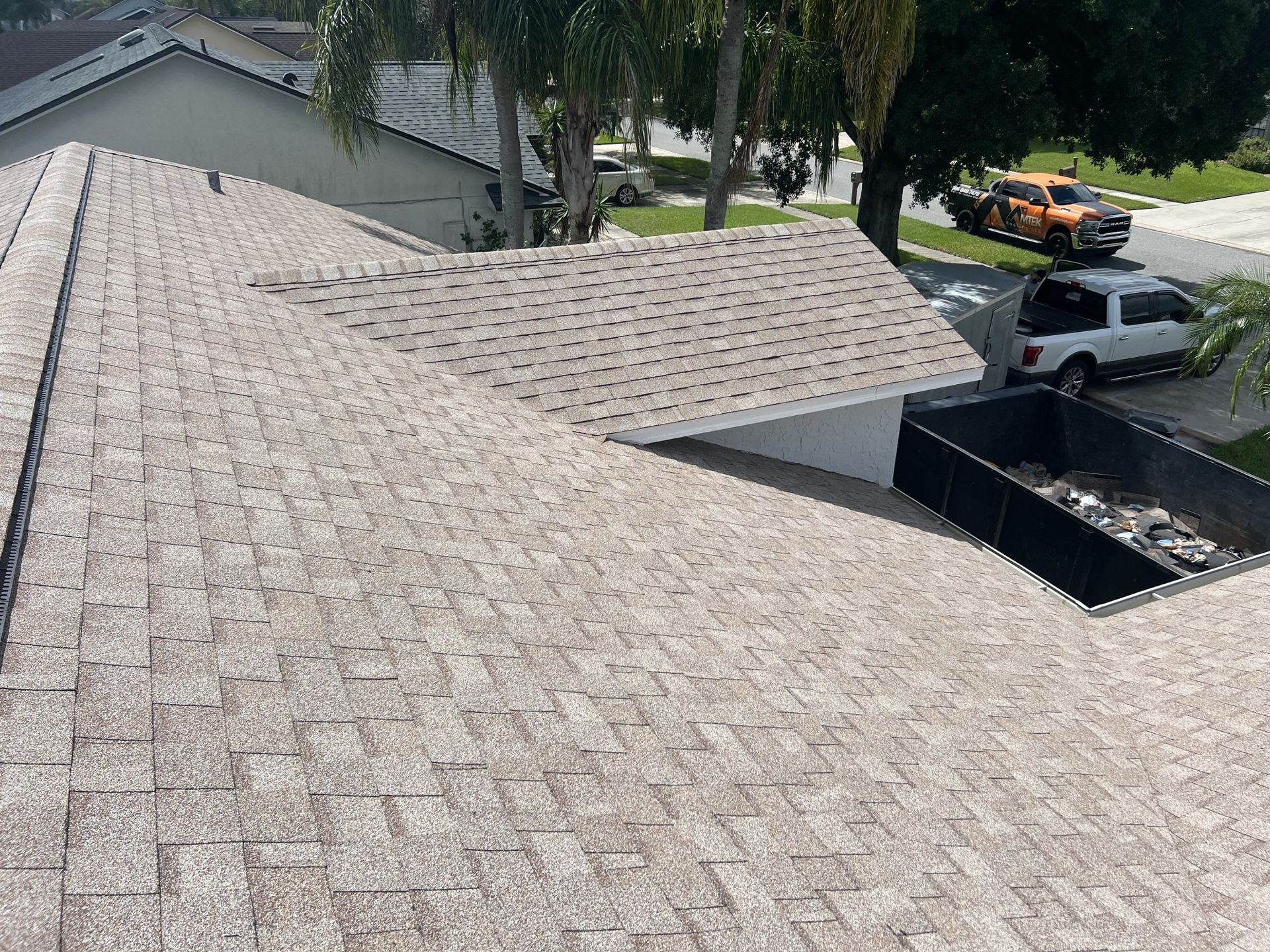 Rooftop view of tan shingle roofing with neighboring houses and parked vehicles below.