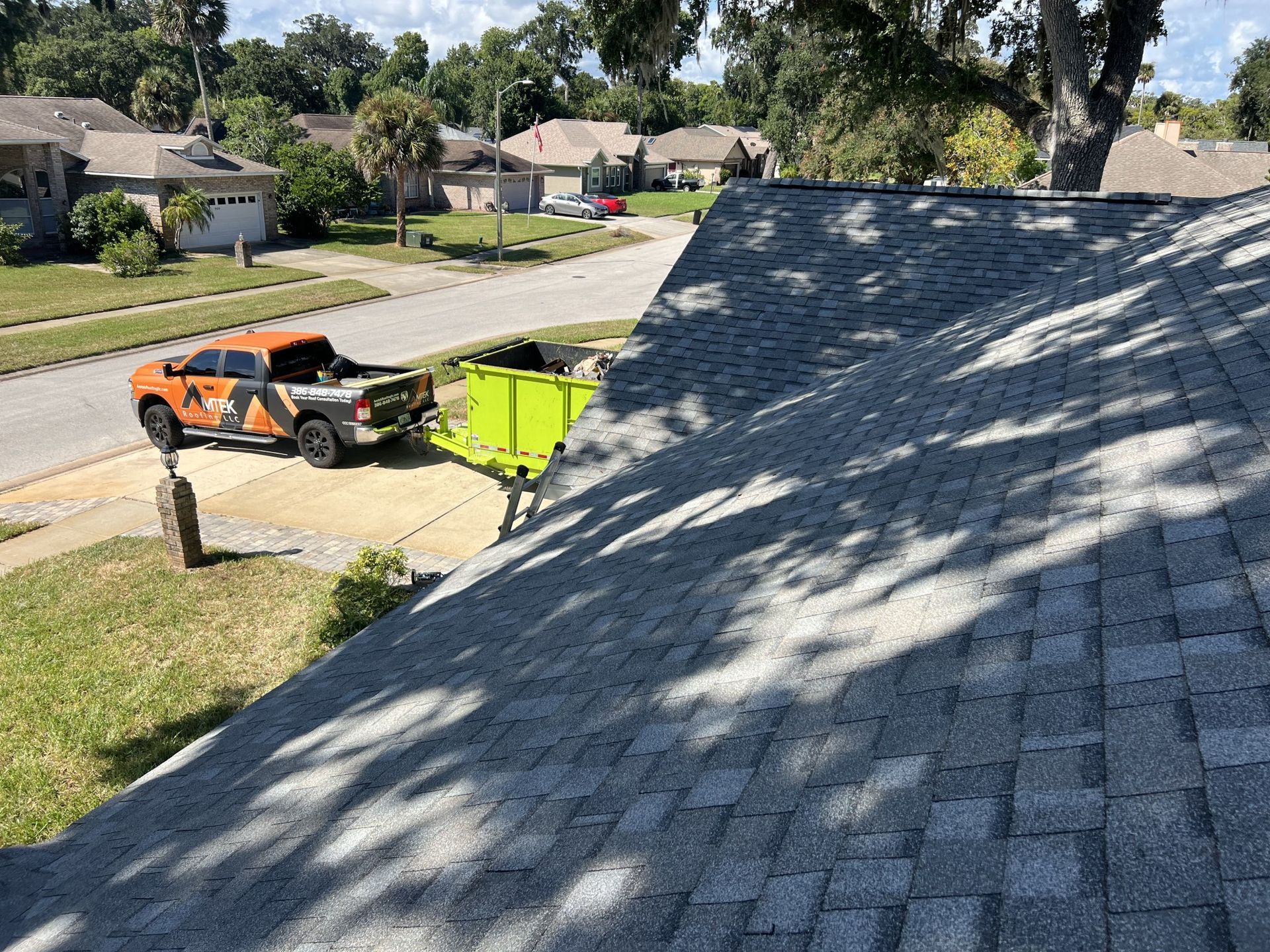 Roof shingles in foreground, with a worker in a truck and lime-green dumpster on a suburban street.