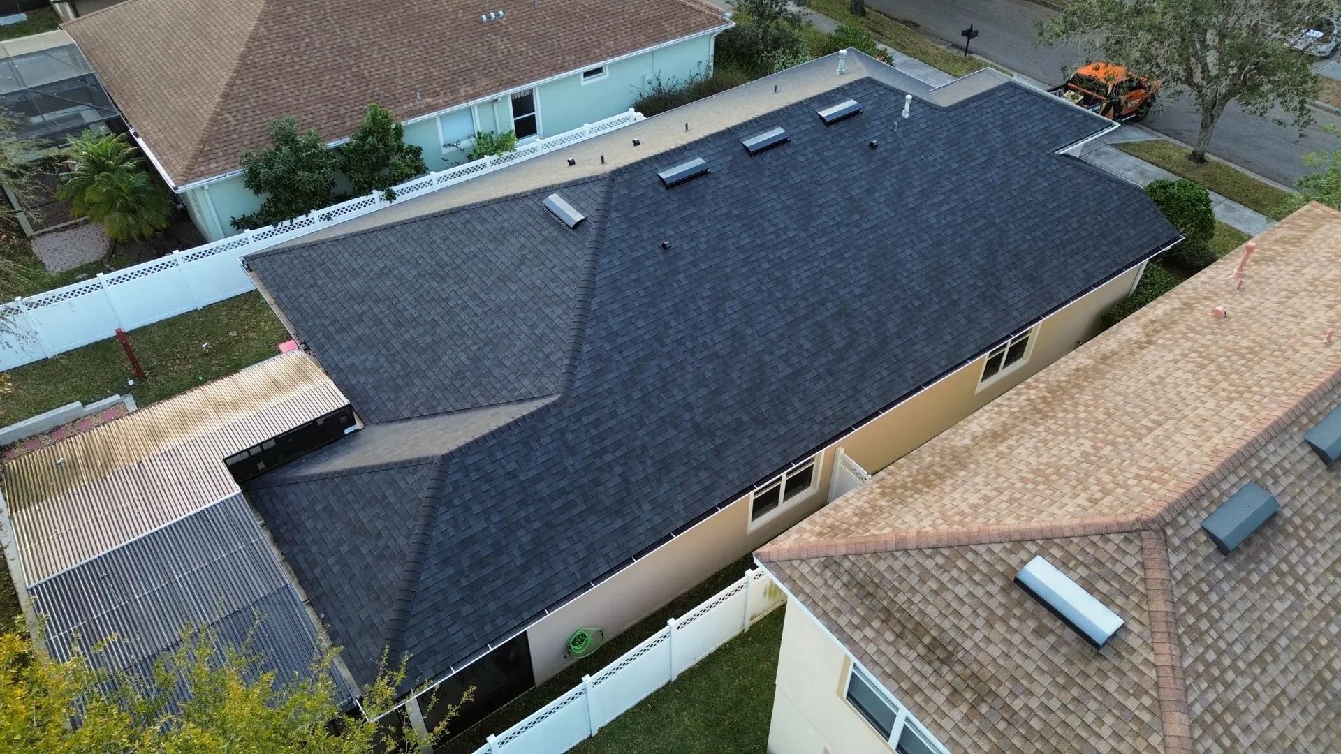 Aerial view of a house with a dark roof, beige walls, and neighboring homes along a residential street.