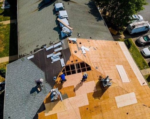 Roofers installing new shingles on a house roof with exposed plywood and gray and orange sections