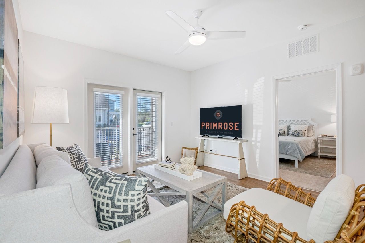 Bright living room in a white-themed apartment with a sofa, coffee table, and TV; doorway to a bedroom.