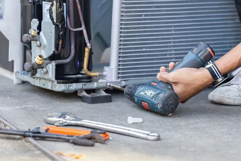 Person using a power drill on an air conditioning unit. Tools are scattered on the ground.