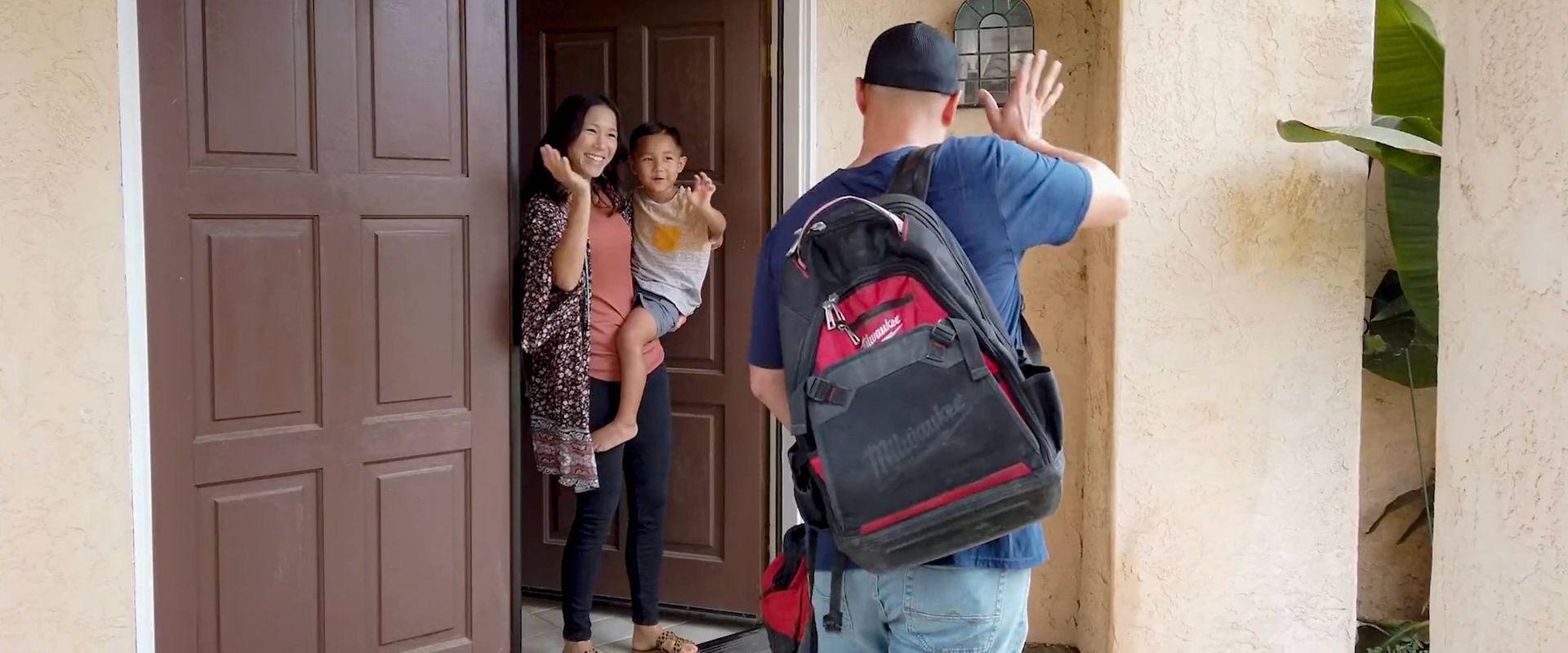 A man with a backpack waves goodbye to a woman and child at a doorway.