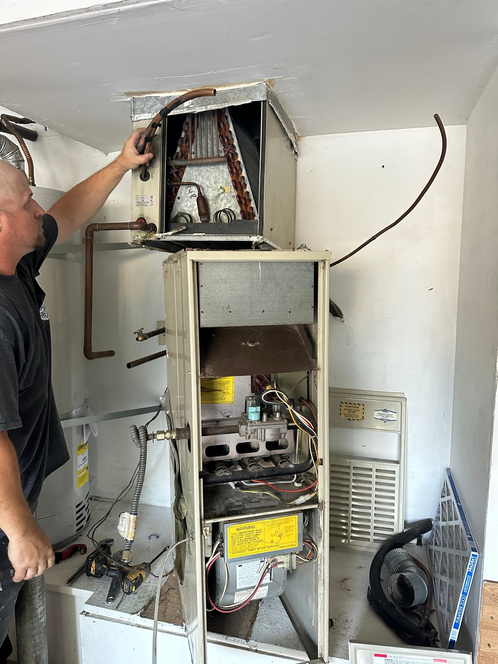 Man inspecting an exposed HVAC unit. White and brown components. Tools visible. In a white room.