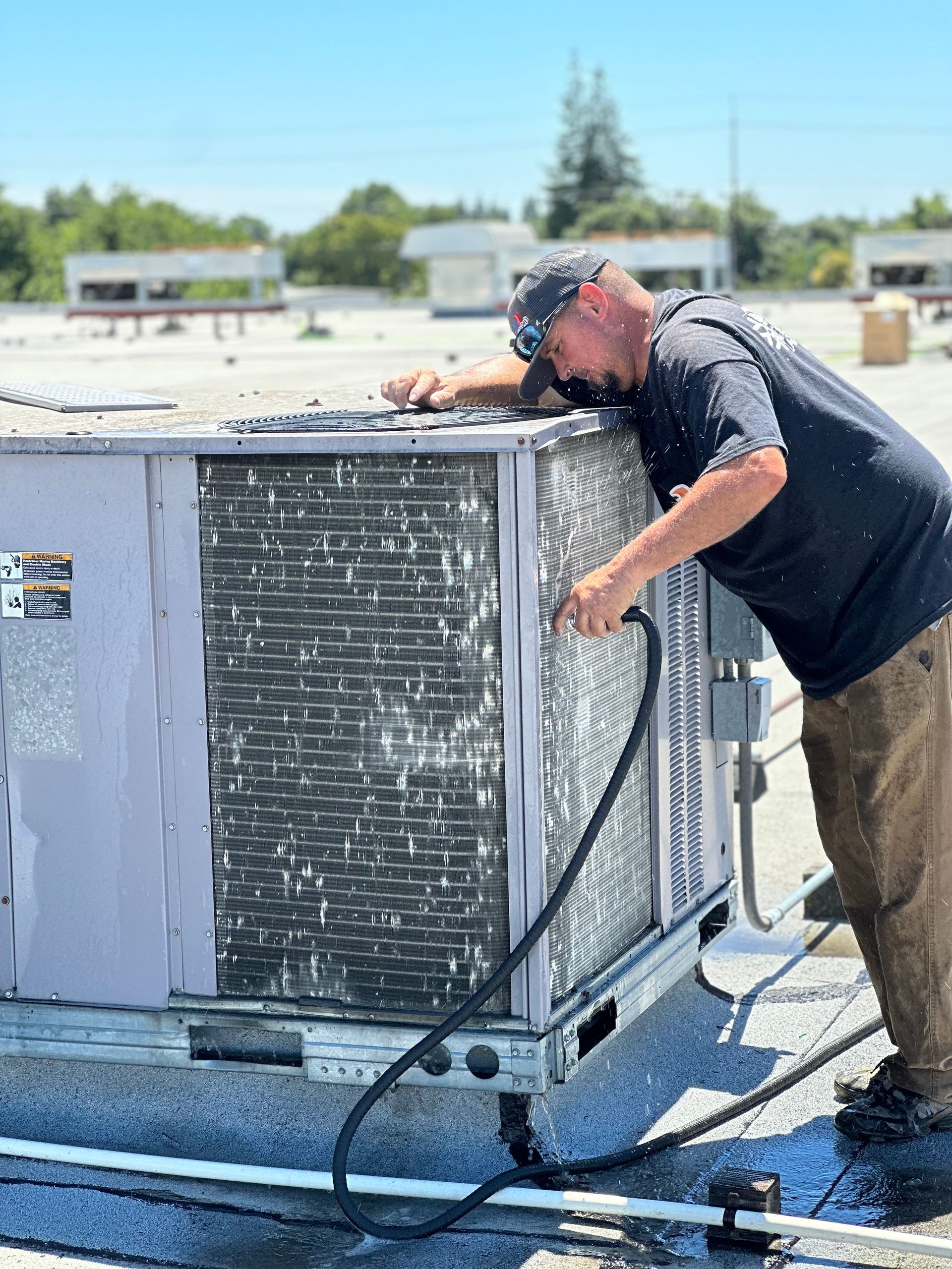 Man cleaning an air conditioning unit on a rooftop with a hose.