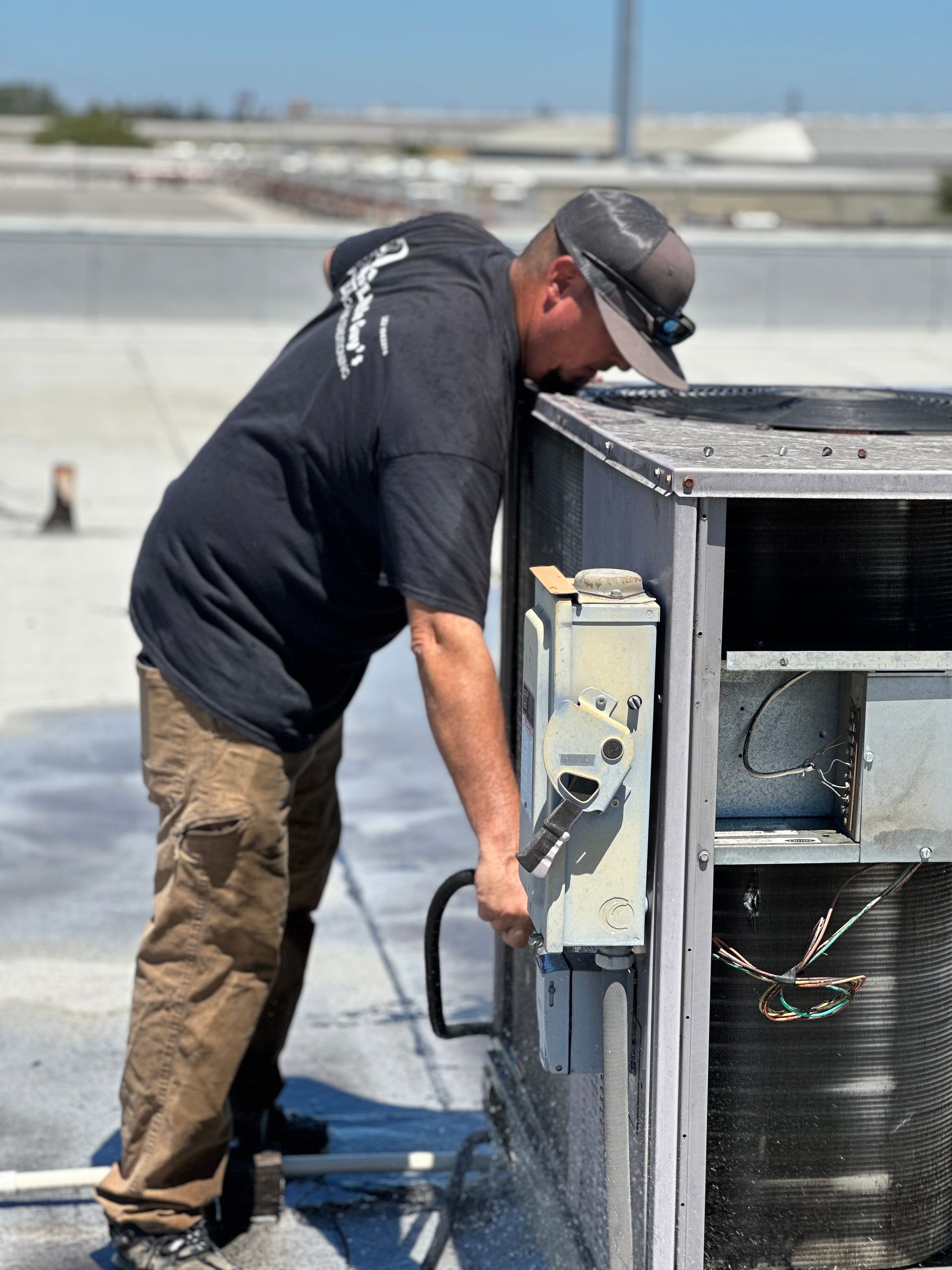 HVAC technician working on rooftop unit, using a wrench. Wearing a black shirt, brown pants, and a baseball cap.