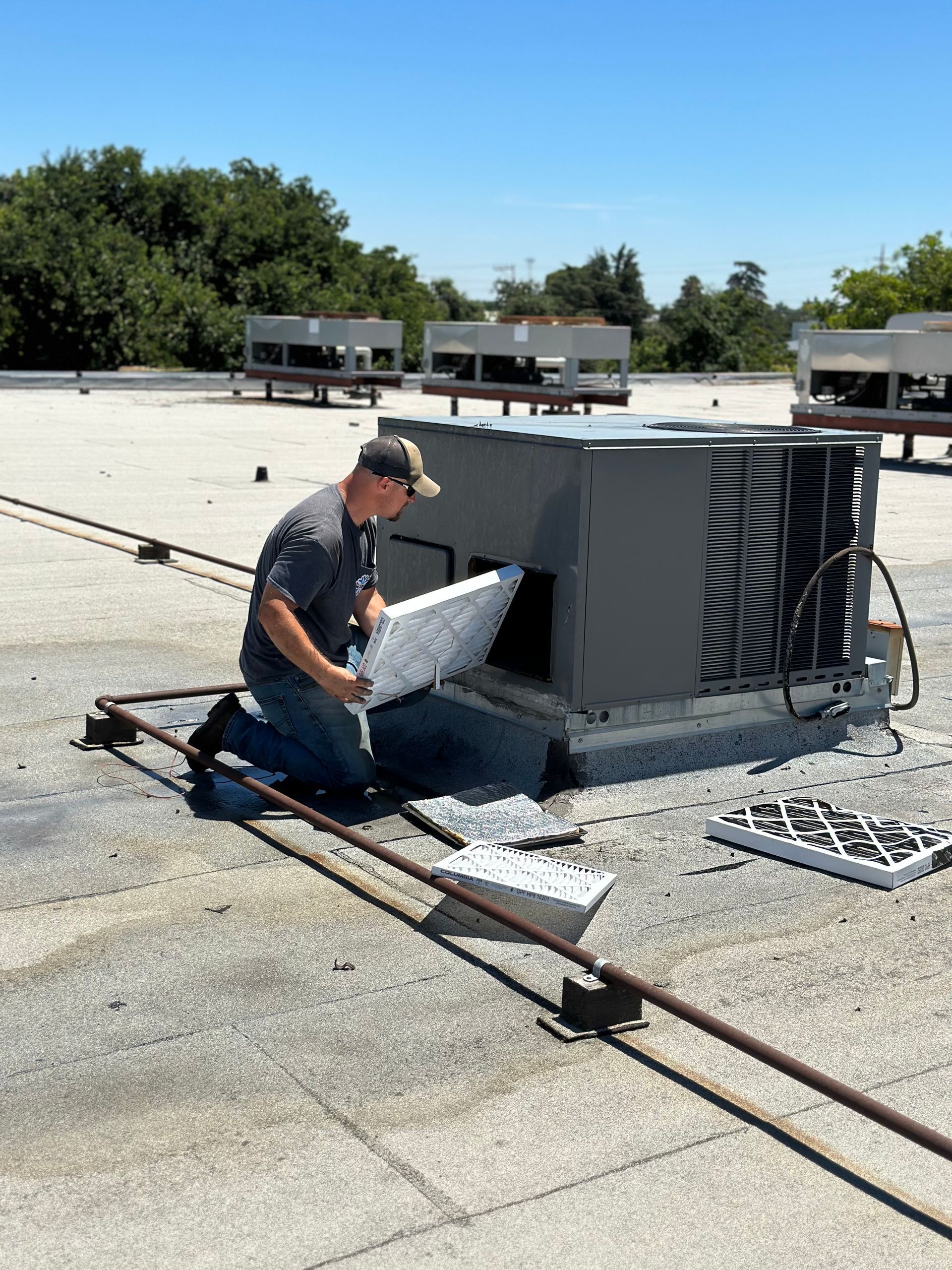 A person kneeling, changing an air filter on a rooftop HVAC unit; sunny day.