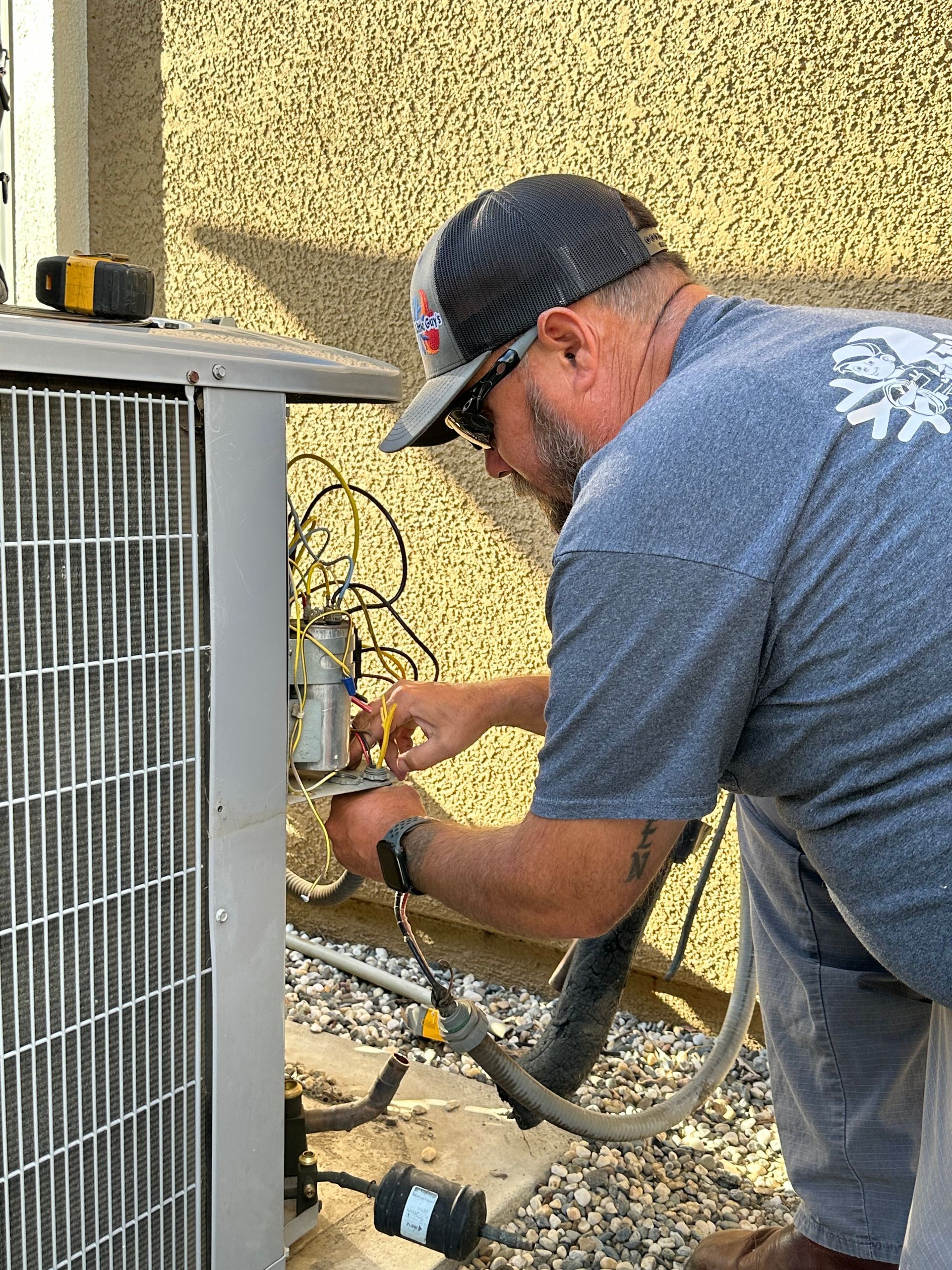 HVAC technician works on an air conditioning unit outside, wearing a hat and sunglasses.