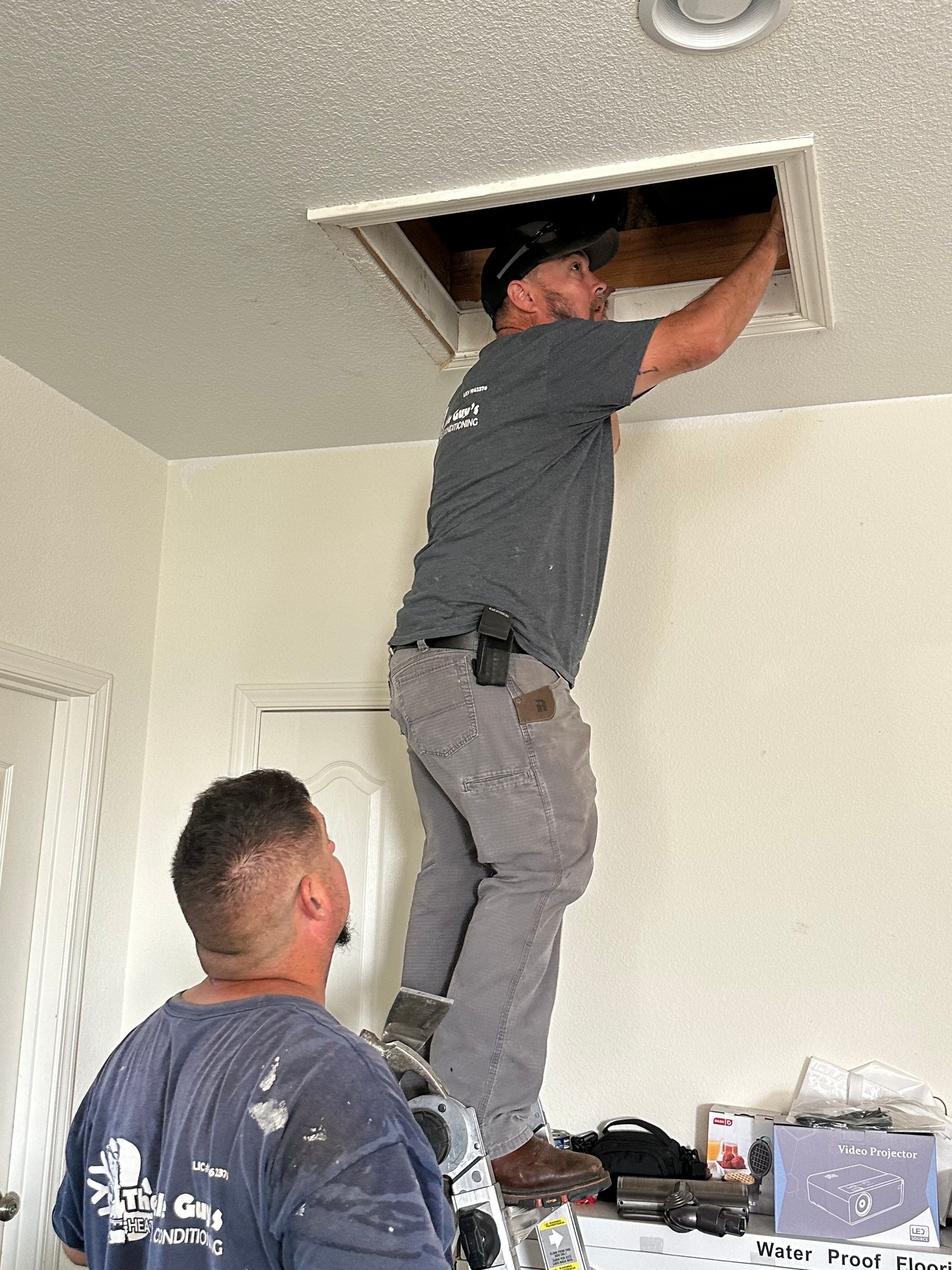 Two men working on an attic access panel in a ceiling. One stands on a ladder, reaching up; another looks up.