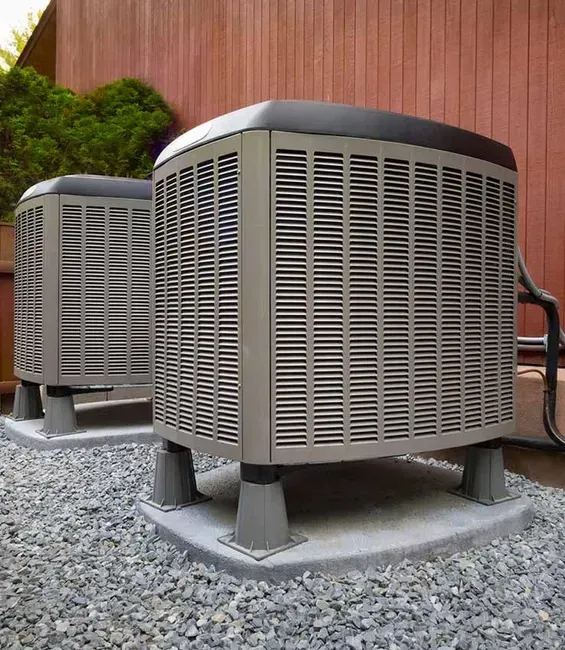 Two gray air conditioning units on concrete pads, gravel ground, wooden fence backdrop.