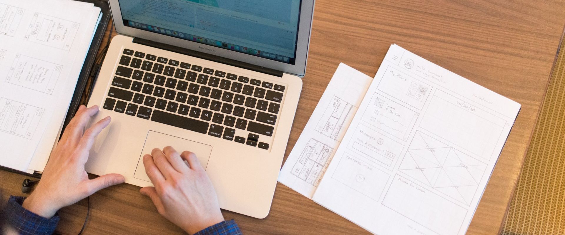 A person using a laptop on a wooden table, with papers nearby.
