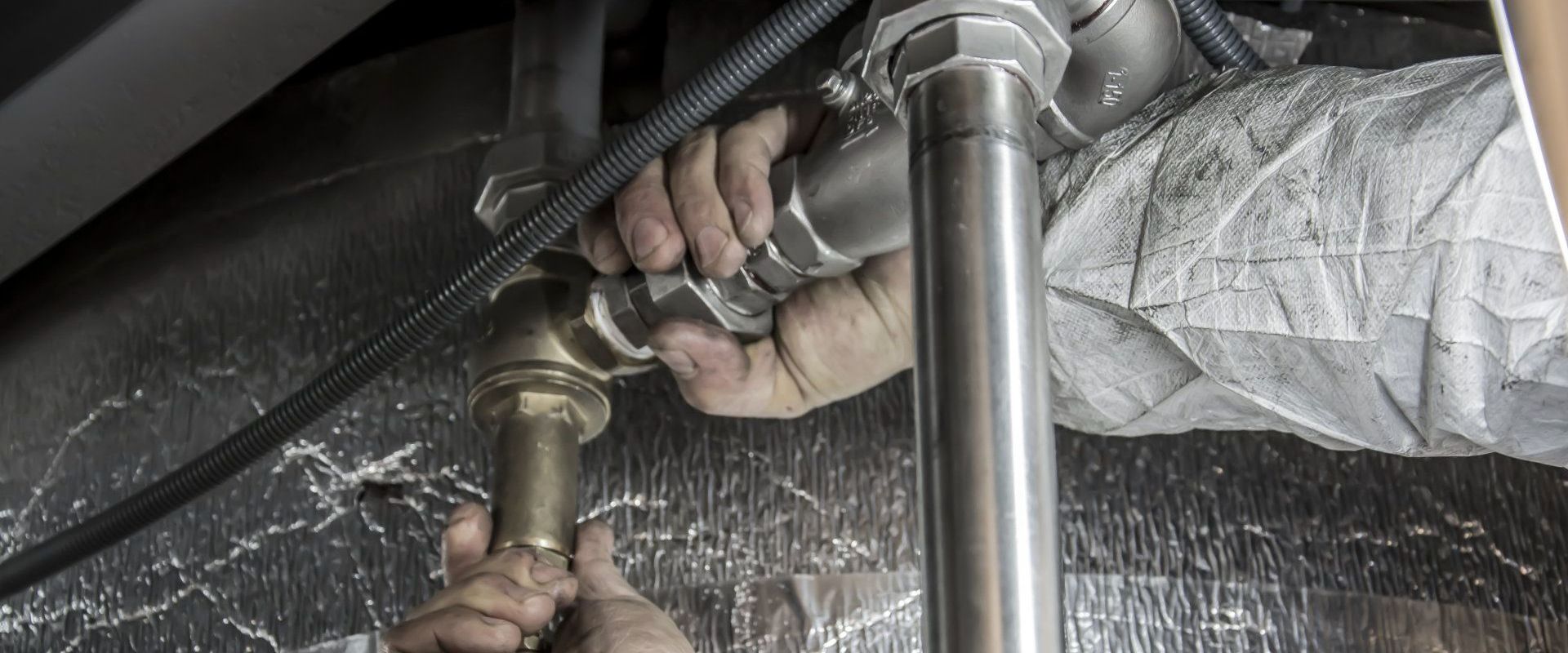 Hands tightening a metal pipe fitting in a confined space.