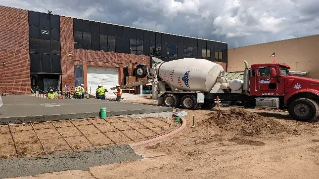 A red concrete mixer truck is driving down a dirt road in front of a building.