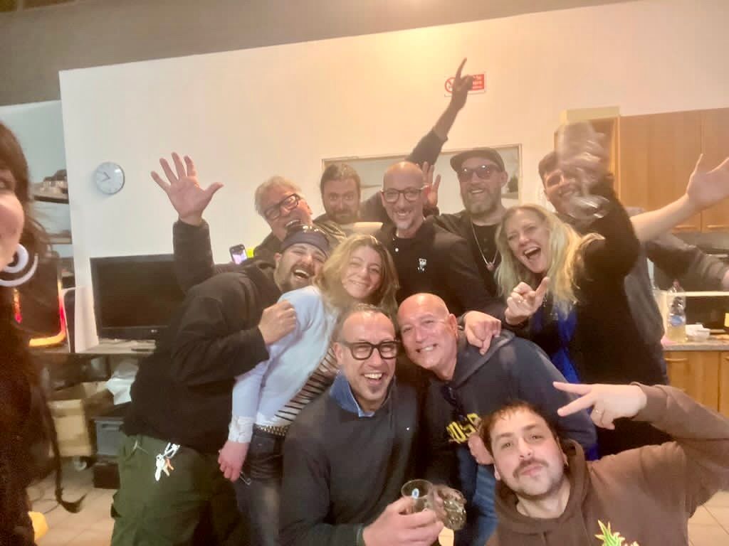 Group of people smiling and waving in an indoor setting. Some are holding up fingers, arms, or cameras.