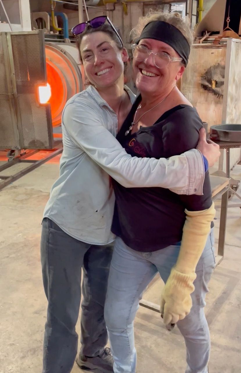 Two people hugging in a glassblowing studio, smiling. One wears a black headband, the other a light blue shirt.