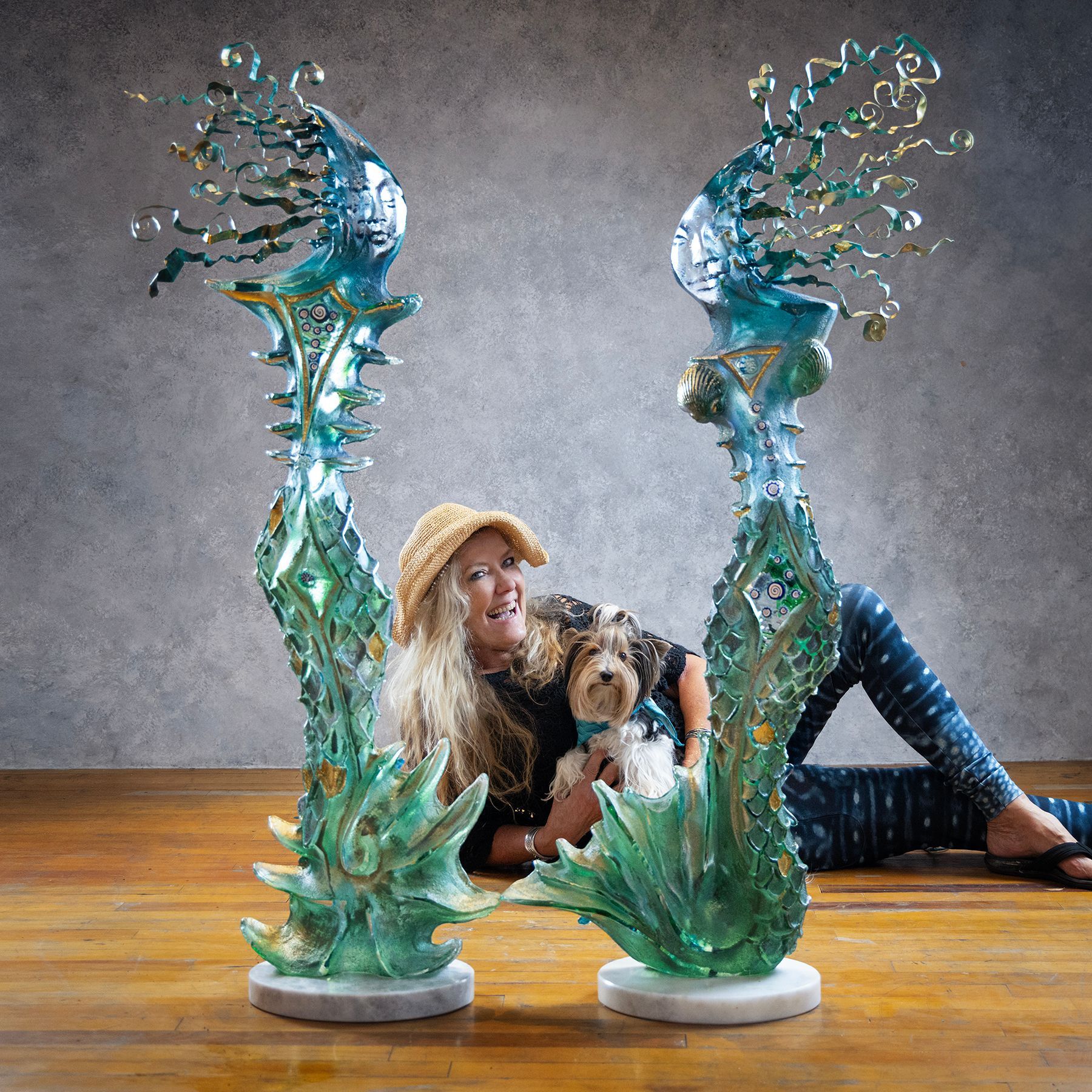 A woman is sitting on the floor holding a dog next to two sculptures.