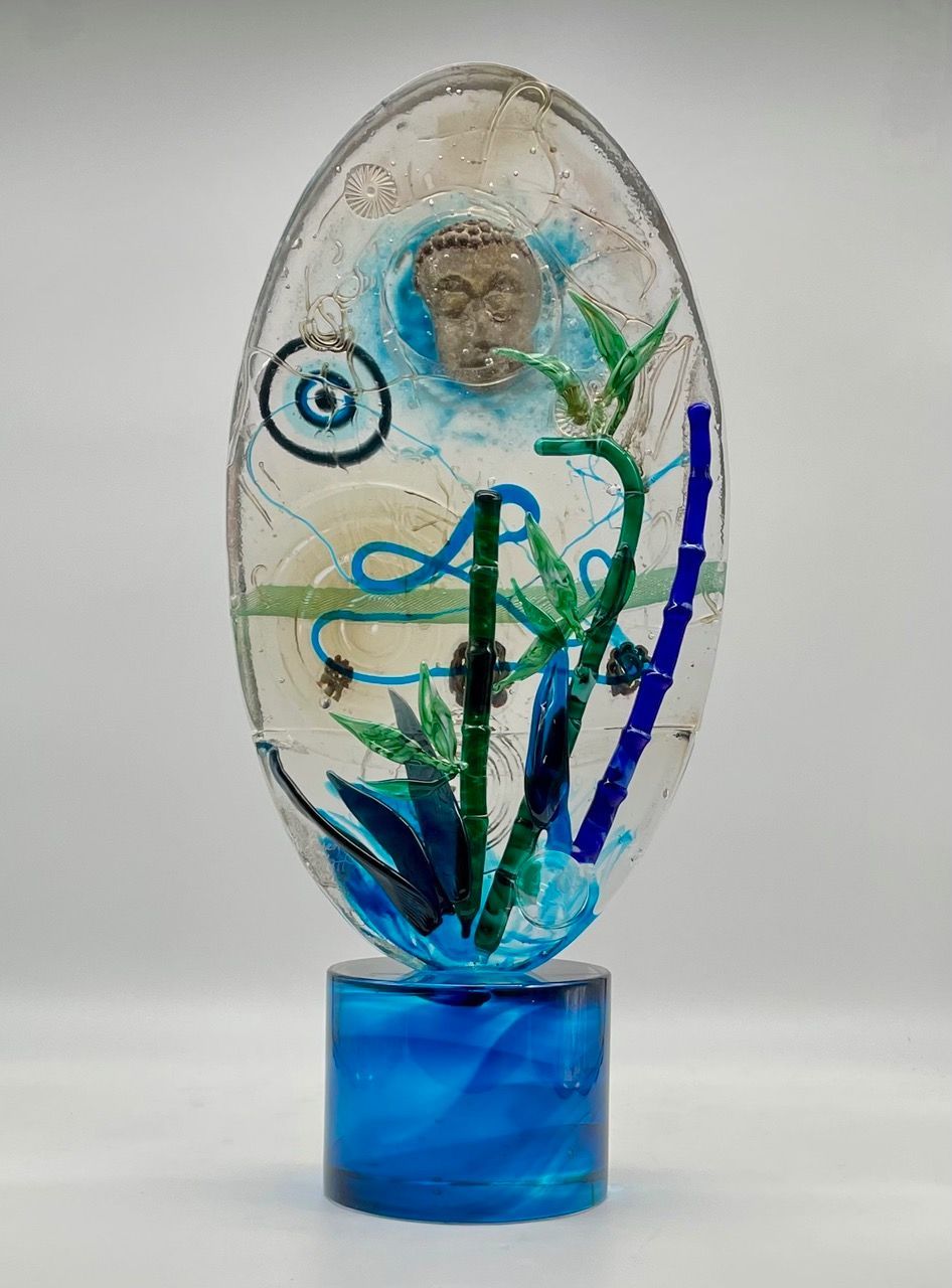 Oval glass sculpture with a Buddha head, bamboo, and blue base.