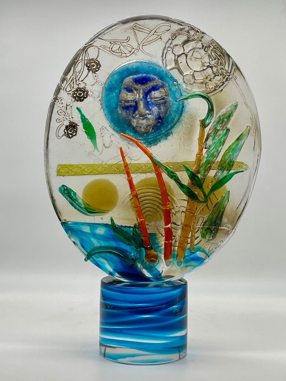 Glass sculpture with blue face, sun, and plants on a blue wave base.