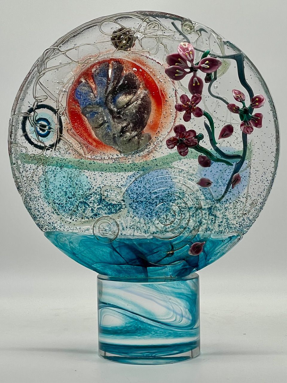 Fused glass sculpture of a landscape with cherry blossoms, blues, and a red sun.