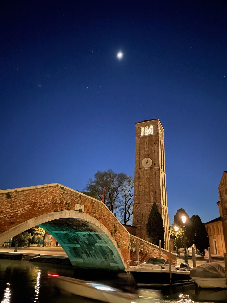 Brick bridge and clock tower at night, with boat in water. Dark blue sky with moon and stars.