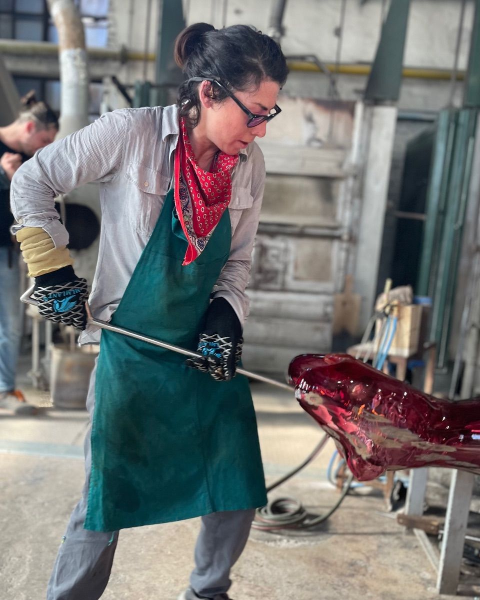 Woman working with molten red glass, using a tool to shape the sculpture. In a workshop, wearing safety gear.
