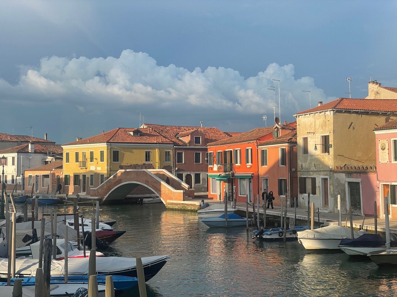 Colorful buildings line a canal in Burano, Italy, with boats docked and a bridge under a cloudy sky.