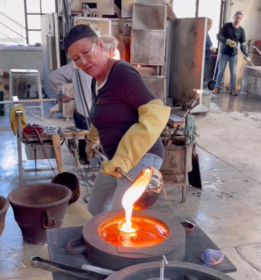 Woman pouring molten glass from a crucible into a mold in a glassblowing studio.