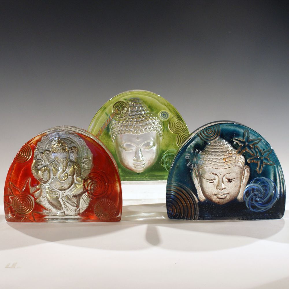 Three arched glass sculptures featuring silver Buddha faces, in red, green, and blue.
