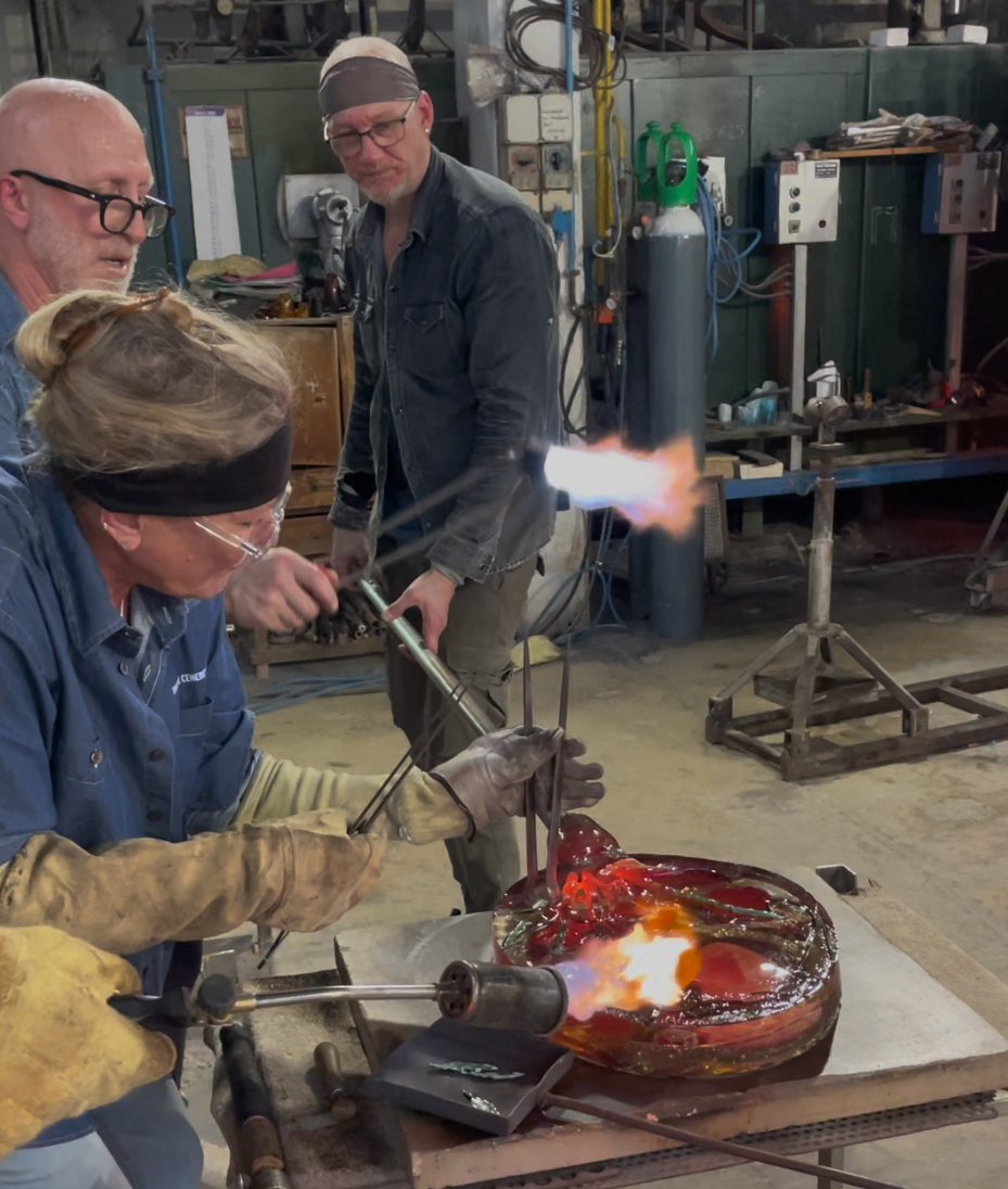 Three people using torches to shape molten glass in a workshop. Flames are bright orange.