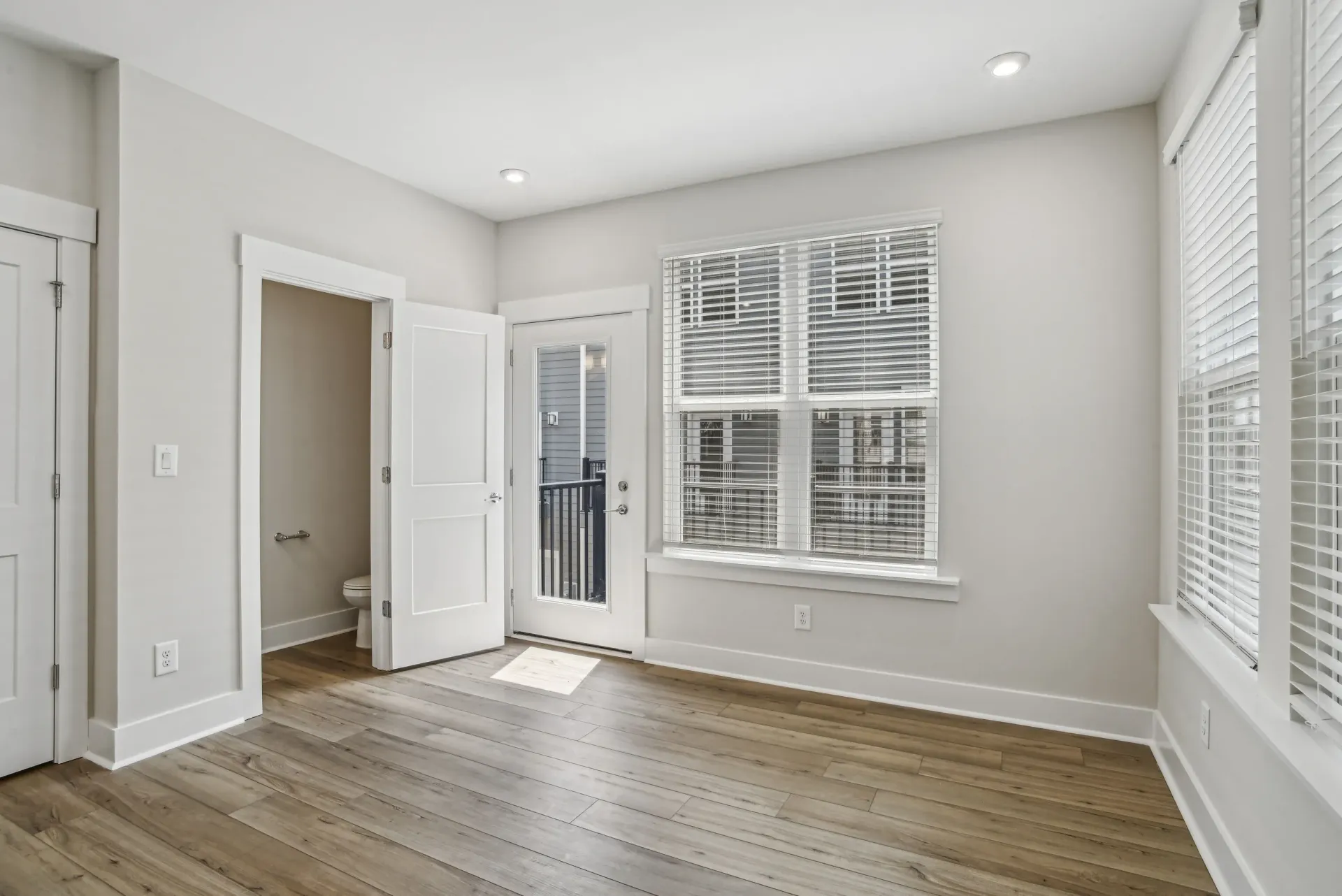 Empty apartment living area with large windows, balcony door, and light wood flooring.