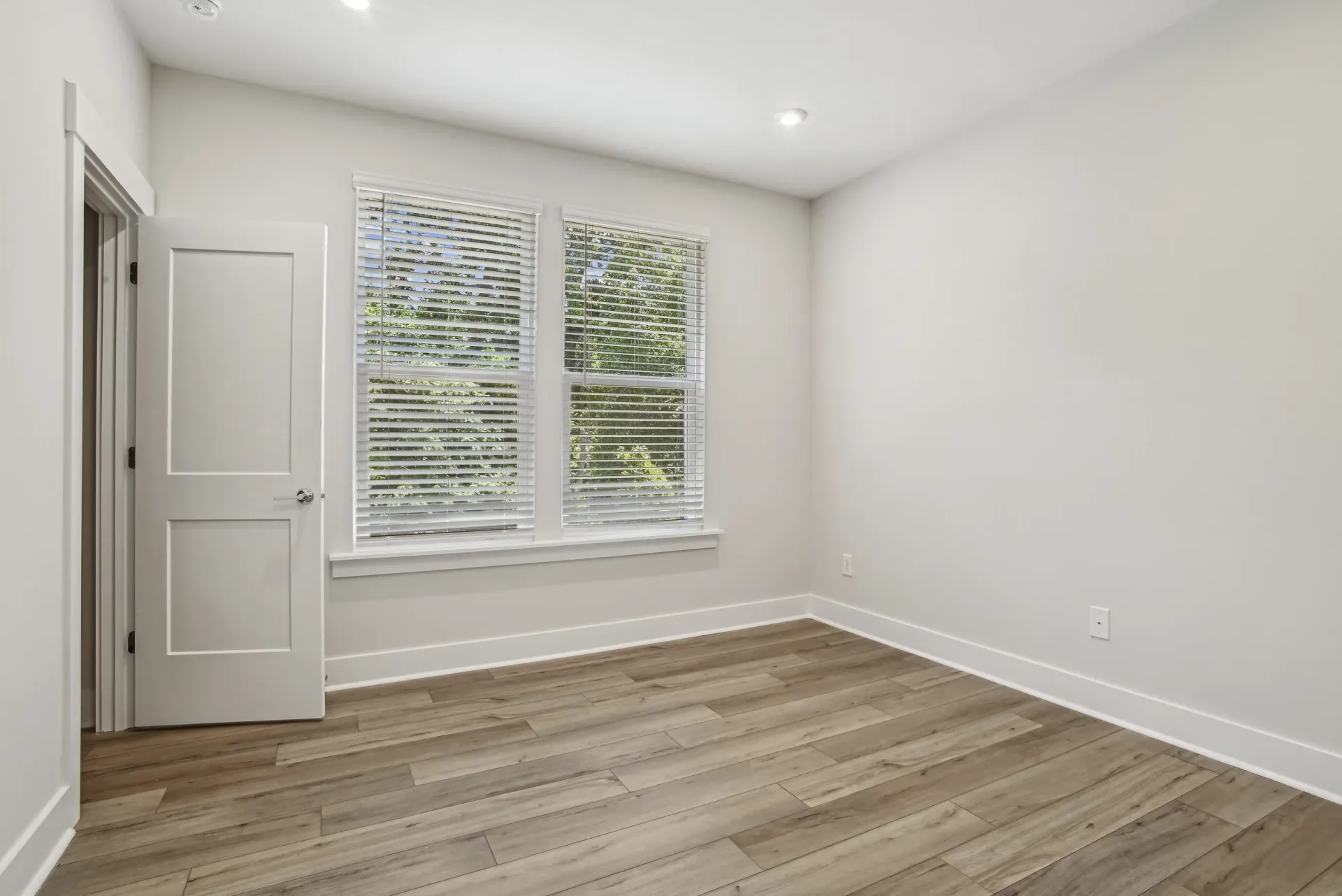 Empty apartment room with two large windows, white blinds, white walls, and wood-look flooring.