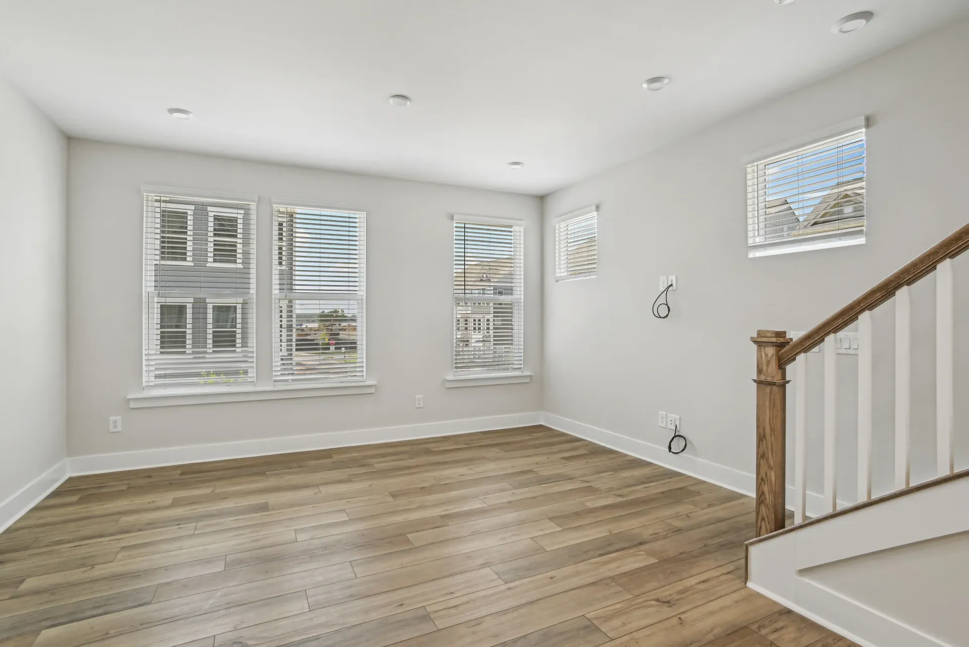 Empty living room in an apartment with wood-look flooring, multiple windows with blinds, and a staircase.