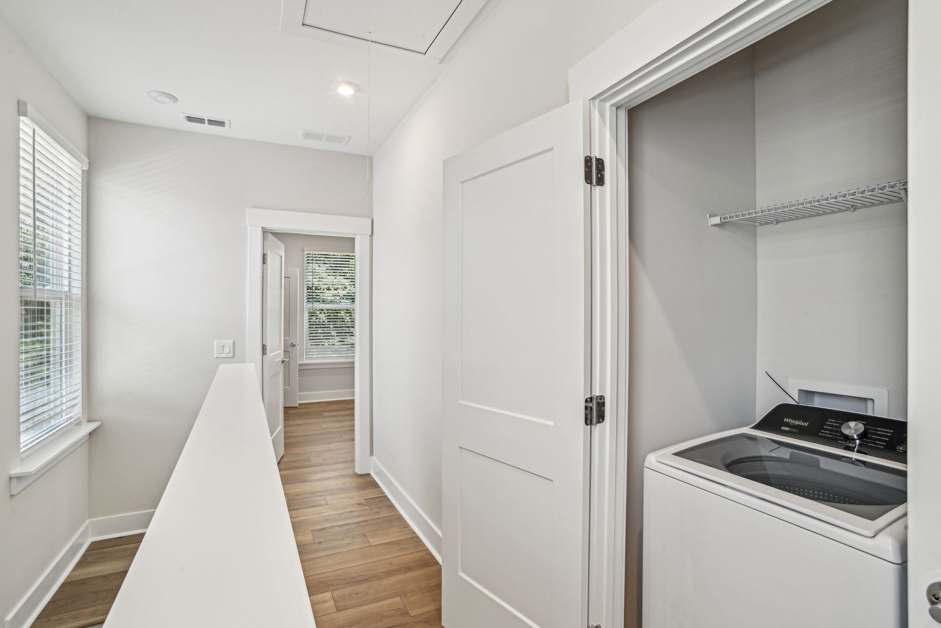 Hallway with laundry closet on the right and window on the left. White walls, wooden floor, and a closed door.